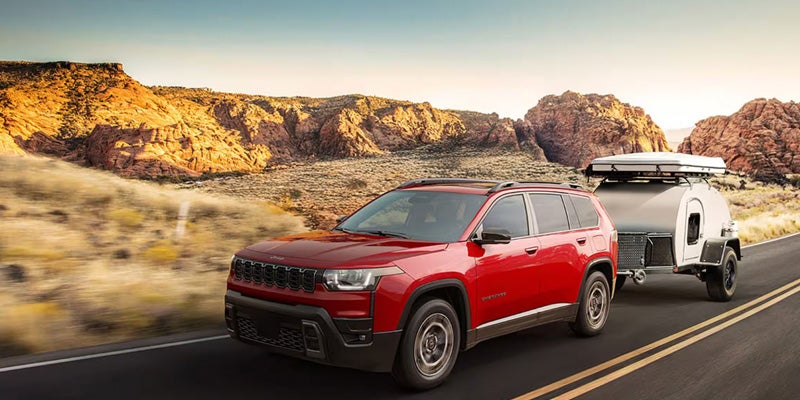 Red Jeep Cherokee towing a small camper trailer on a desert highway surrounded by rocky landscapes.